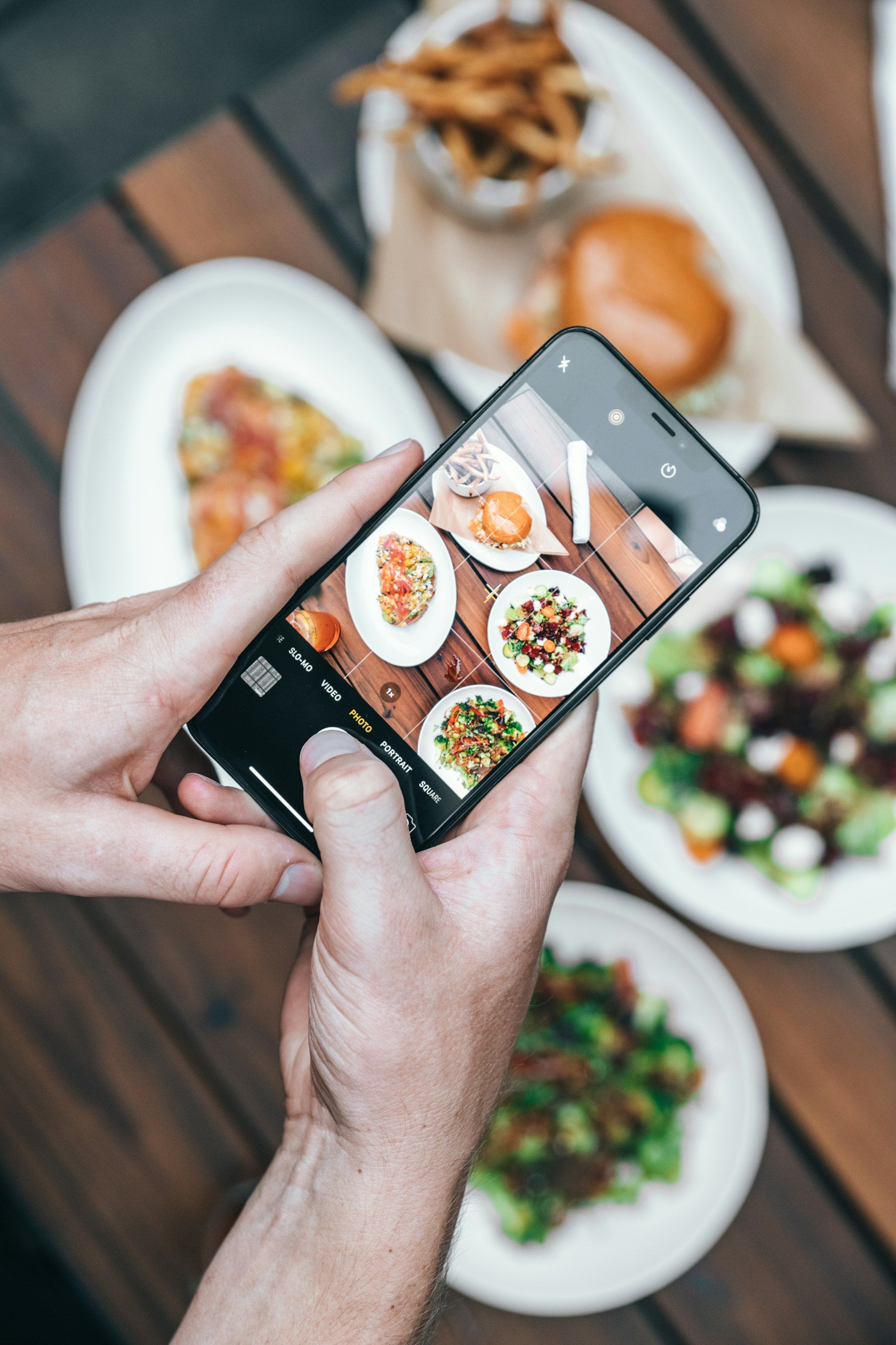 Hände halten ein Smartphone über einen gedeckten Tisch mit Burger, Pommes und Salaten